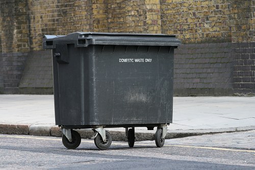 A large black wheeled rubbish bin positioned on the pavement in an urban area, with its lid closed and marked with the words 'DOMESTIC WASTE ONLY' in white lettering. The bin is made of plastic with a textured surface and is situated near a brick wall with visible weathering and a concrete retaining wall behind it. The surrounding pavement is light grey, and the curb is slightly raised with a small section of road visible to the left. This scene reflects a typical outdoor rubbish collection point, relevant to commercial waste management services such as those provided by Commercial Waste Penge in the Penge area. The lighting indicates a cloudy day, with diffuse illumination highlighting the bin’s surface and the nearby environment, emphasizing the importance of proper waste disposal infrastructure in urban settings.