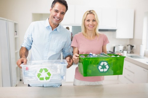A smiling man and woman standing in a modern kitchen, each holding a transparent recycling bin with a prominent green recycling symbol on the front. The man's bin is empty and made of clear plastic, while the woman's bin is a bright green plastic container filled with recyclable materials. They are positioned in the foreground, with kitchen cabinets, a stove, and various kitchen utensils visible in the background, suggesting a domestic environment. The image emphasizes environmentally responsible waste disposal and recycling practices, which aligns with services provided by Commercial Waste Penge for proper rubbish management and collection in the local area. The bright lighting and clean setting highlight the importance of organized waste separation in residential properties, subtly supporting the importance of professional rubbish removal services in the Penge postcode area. Overall, the image underscores the community's commitment to recycling and waste reduction at home, consistent with the company's focus on waste management solutions for domestic customers in the region.