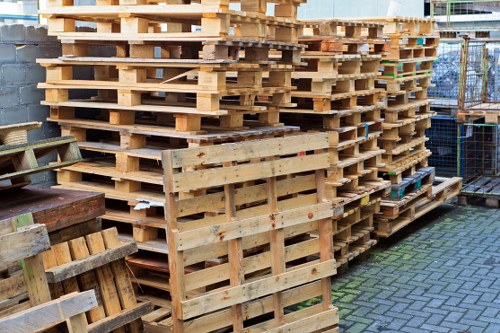 A large stack of light-colored wooden pallets arranged in multiple columns and tiers, situated outdoors on a paved surface with interlocking bricks. The pallets have a rough, unfinished texture and are stacked neatly, some showing signs of usage such as slight stains or minor wear. Surrounding the pallets are additional wooden items and metal fencing, indicating a storage or waste management area in a commercial setting. The background includes partial views of a brick wall and possibly construction or industrial equipment, with natural daylight illuminating the scene to highlight the materials. This image relates to rubbish removal services provided by Commercial Waste Penge, demonstrating the type of bulk waste such as pallets collected and managed in Penge or nearby London areas.
