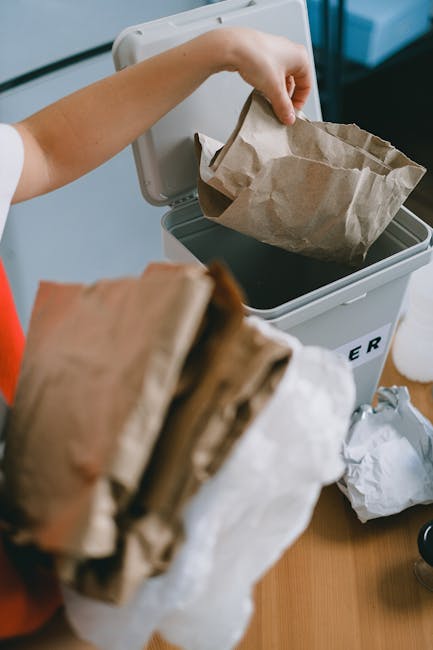 A small, vintage blue flatbed truck parked on the side of a road, loaded with a large amount of mixed waste materials including cardboard boxes, black plastic bags, and various discarded items. The tr